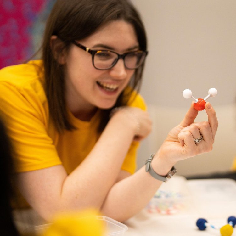 A woman in a yellow t-shirt and glasses shows a model of a molecule to two children