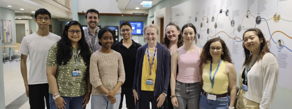 Photo of the Machesky Lab. A group of seven women and two men stand around Prof Machesky. They are all smiling and look happy.