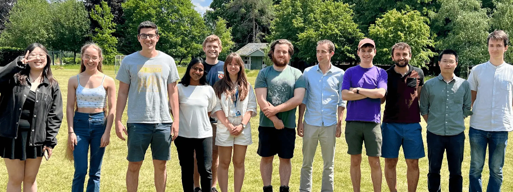 A photo of twelve people standing in a line smiling at the camera. They are in a sunny place surrounded by grass and trees and look relaxed.