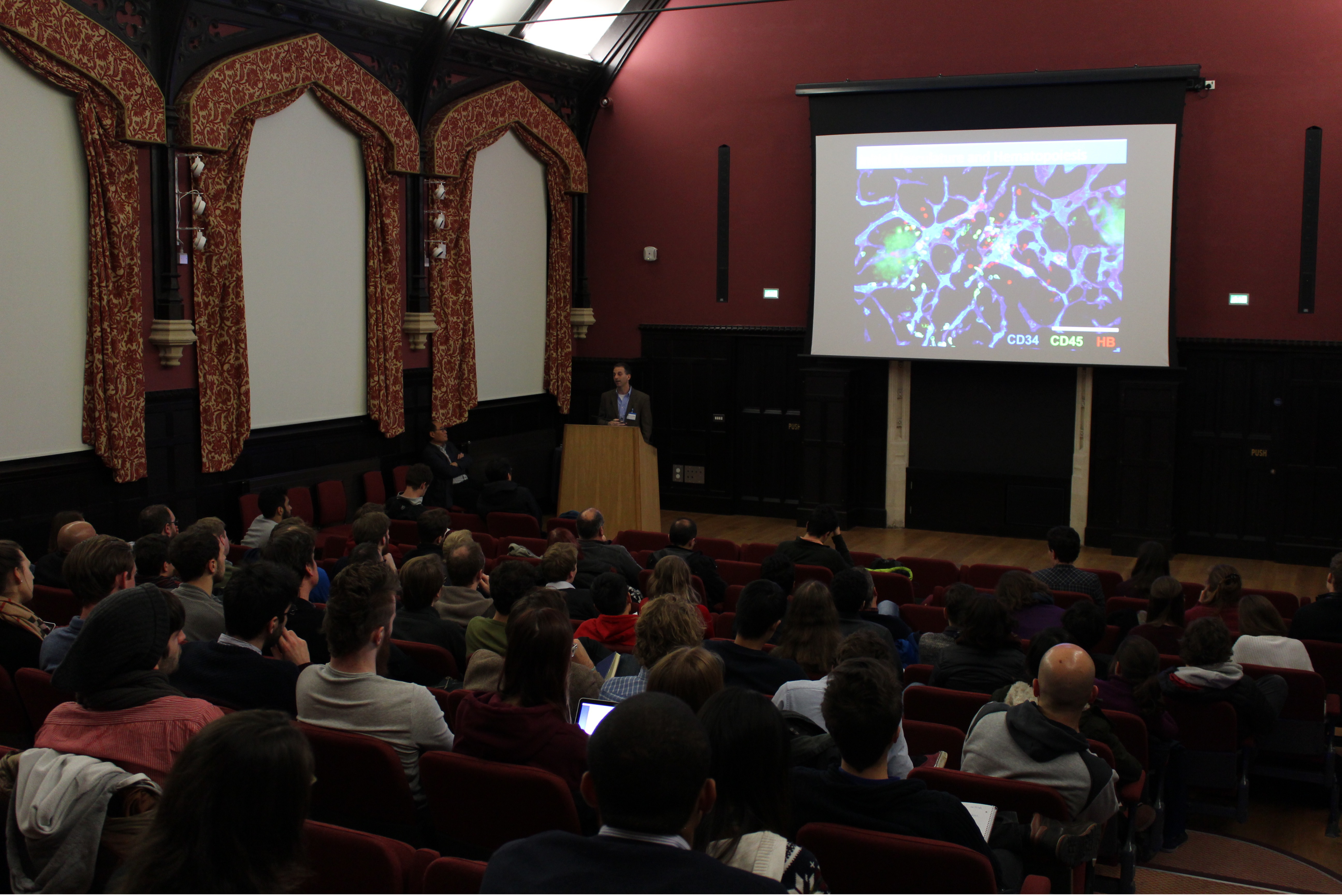 A group of people sit in a darkened auditorium watching a lecture. The building is old and the ceiling is vaulted