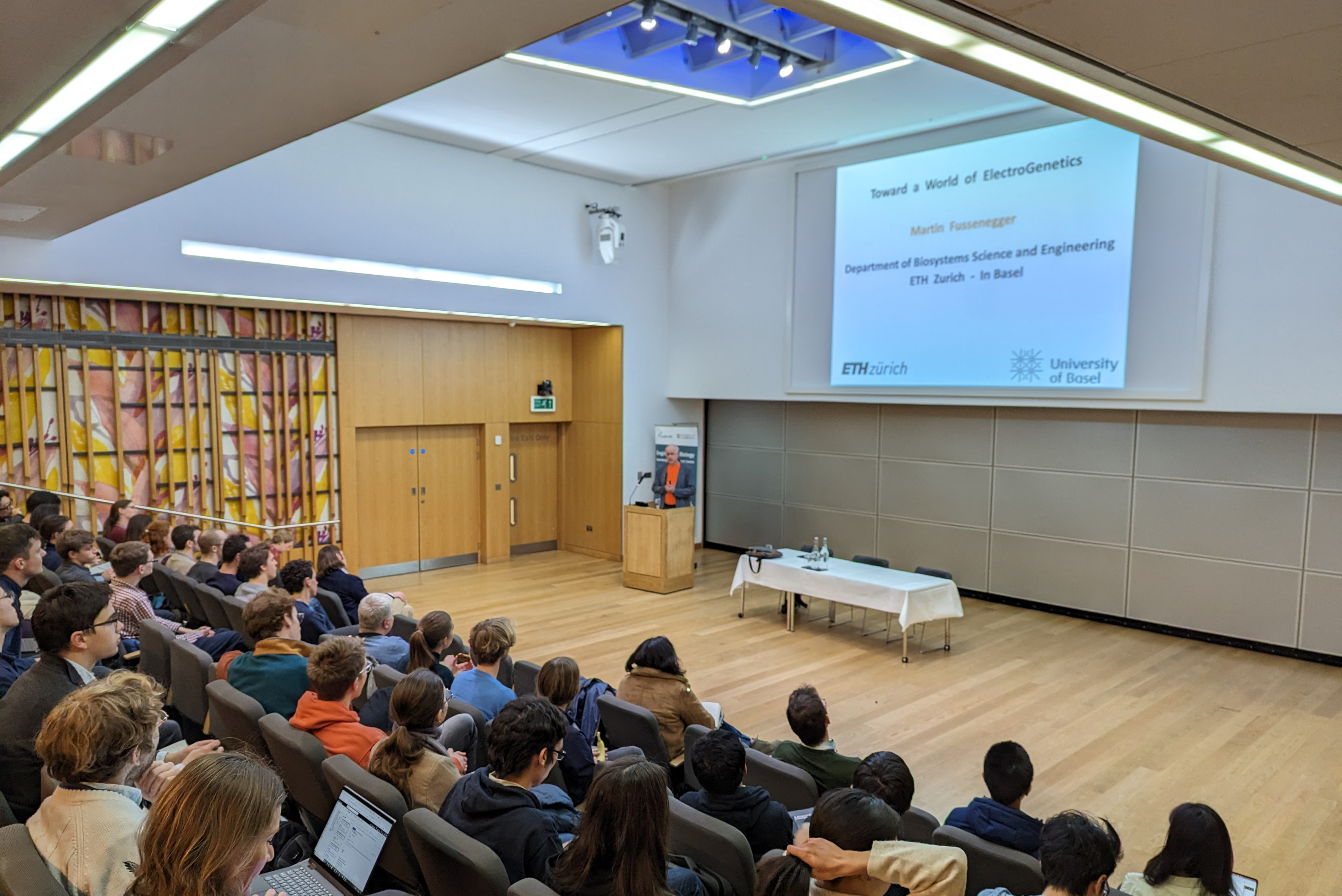 A group of people sit in an auditorium watching a lecture. The building is bright and modern