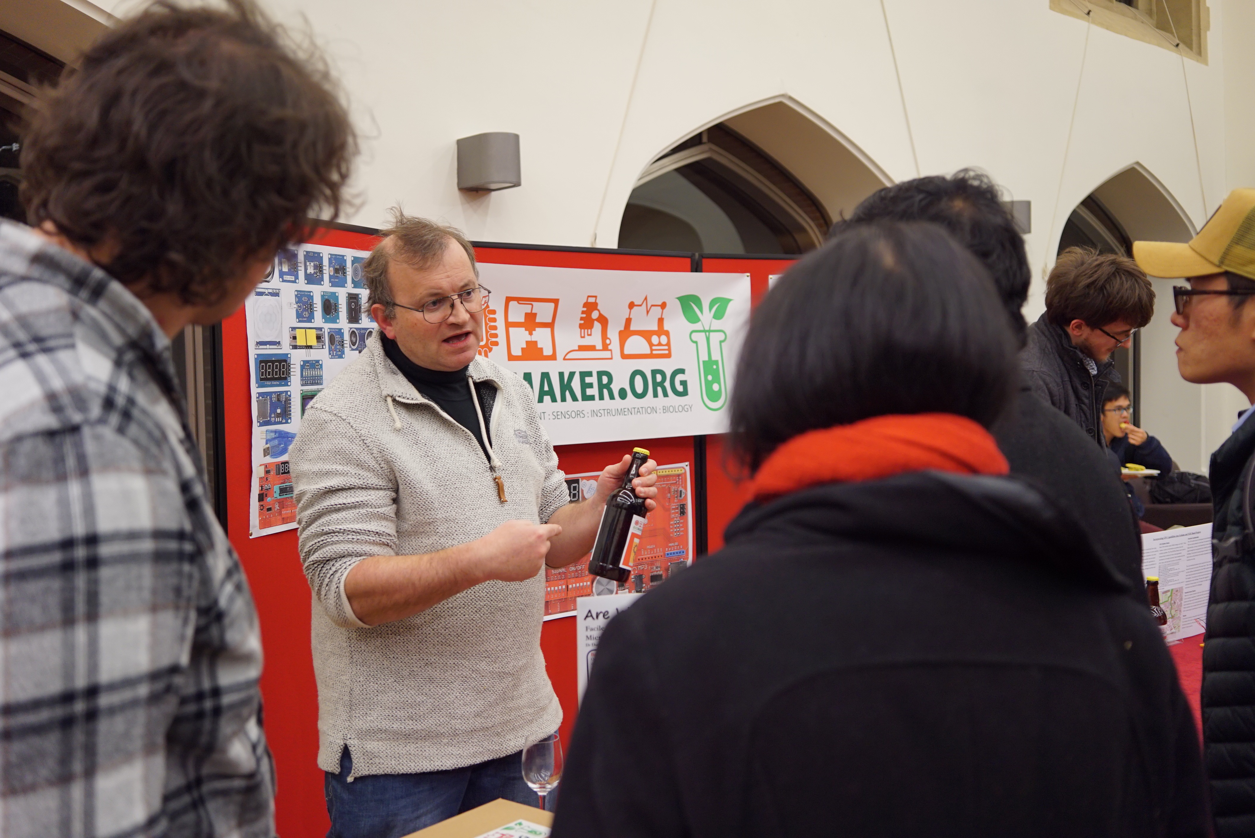 A man stands in front of a poster board with Biomaker branding. He is talking to four people who are facing away from the camera