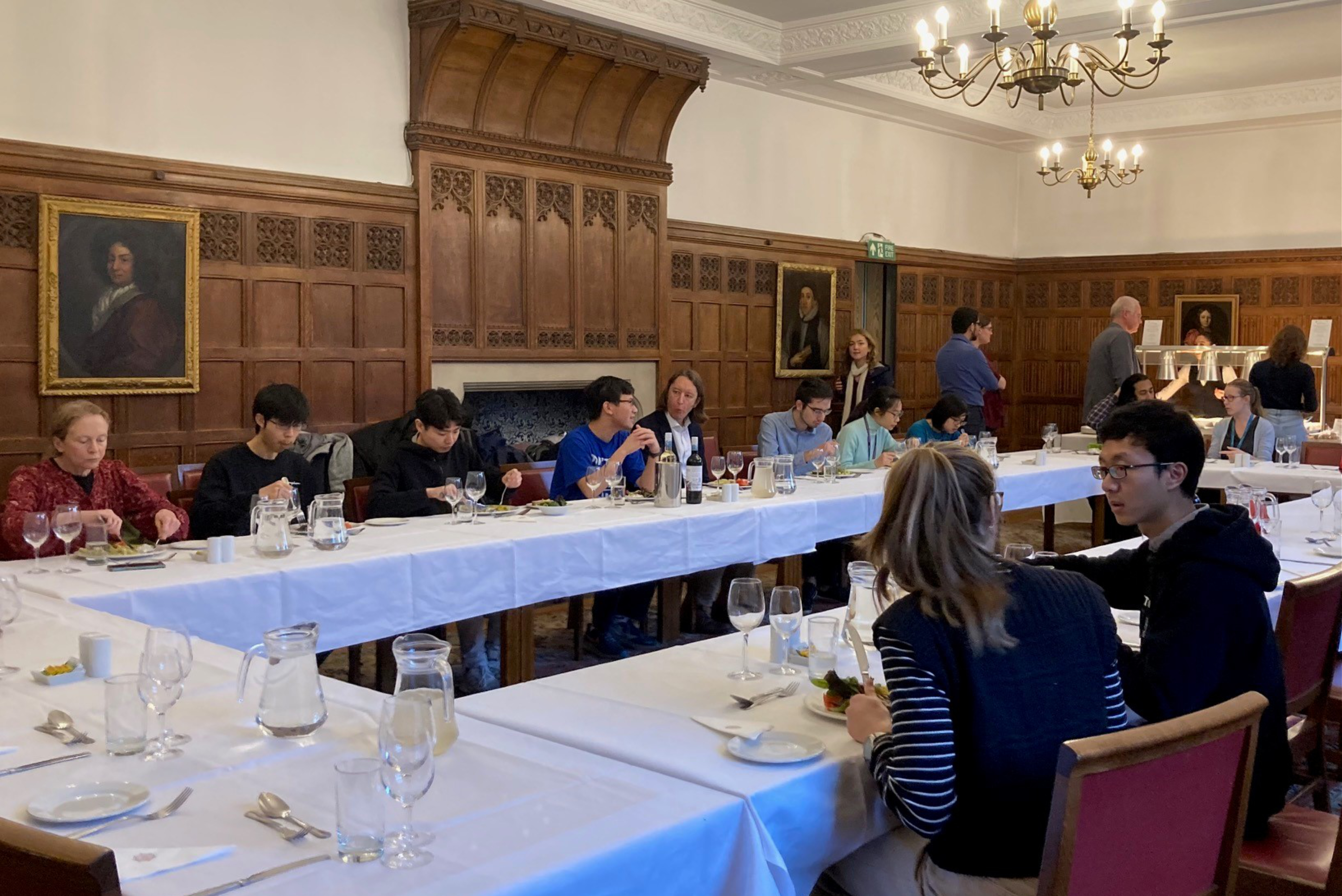 A group of people are eating lunch around a large circular table. They are chatting with each other. The room is old and has wood panelling and old portraits on the walls.