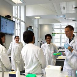 A group of scientists in white lab coats chat and smile at the bench