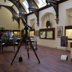 A photograph of a room inside the Whipple museum. It is an old building with wooden floors and a vaulted ceiling. Scientific instruments can been seen on the left, including an old telescope, and desk with a woman sat behind it is on the right/