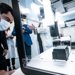 Photo of a man in a suit leaning over an OpenTron lab automation robot. The robot is using a multi-pipette to dispense liquid.