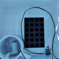 Photo of a plant growth chamber prototype: a white box with a strip of LED lights around the top, inside is a rack for holding ependorph tubes, a mug with tubing coming out and a small electronic sensor