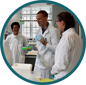 A photo of group of students in lab coats standing by a lab bench and laughing together.