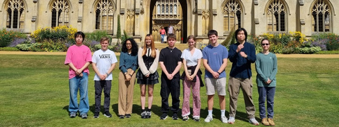 A photo of a group of nine students stood on the grass in front of an ornate gothic building. The students are facing the camera and smiling.