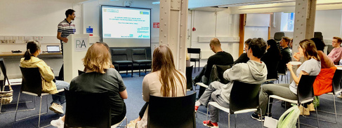 Photo of a group of people sat watching a young man give a presentation