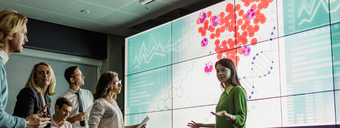 A young woman stands in front of a screen showing a strand of DNA, she is having an informal discussion with a group of people sat in front of her. 