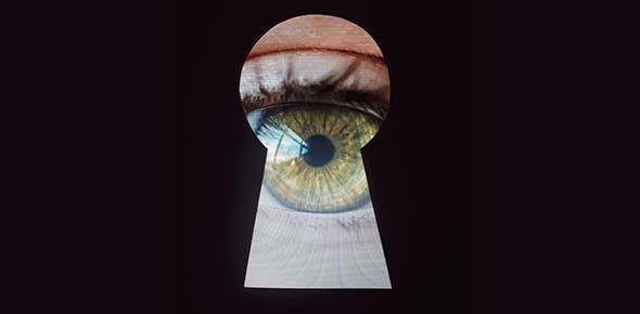 Stock photo of an eye looking through a keyhole. A black background masks a keyhole shape with a green eye peering through.