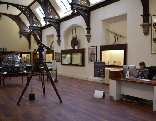 A photograph of a room inside the Whipple museum. It is an old building with wooden floors and a vaulted ceiling. Scientific instruments can been seen on the left, including an old telescope, and desk with a woman sat behind it is on the right/