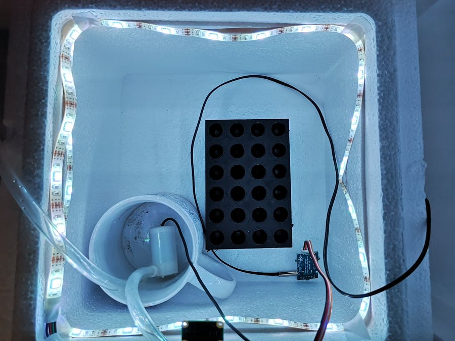 Photo of a plant growth chamber prototype: a white box with a strip of LED lights around the top, inside is a rack for holding ependorph tubes, a mug with tubing coming out and a small electronic sensor